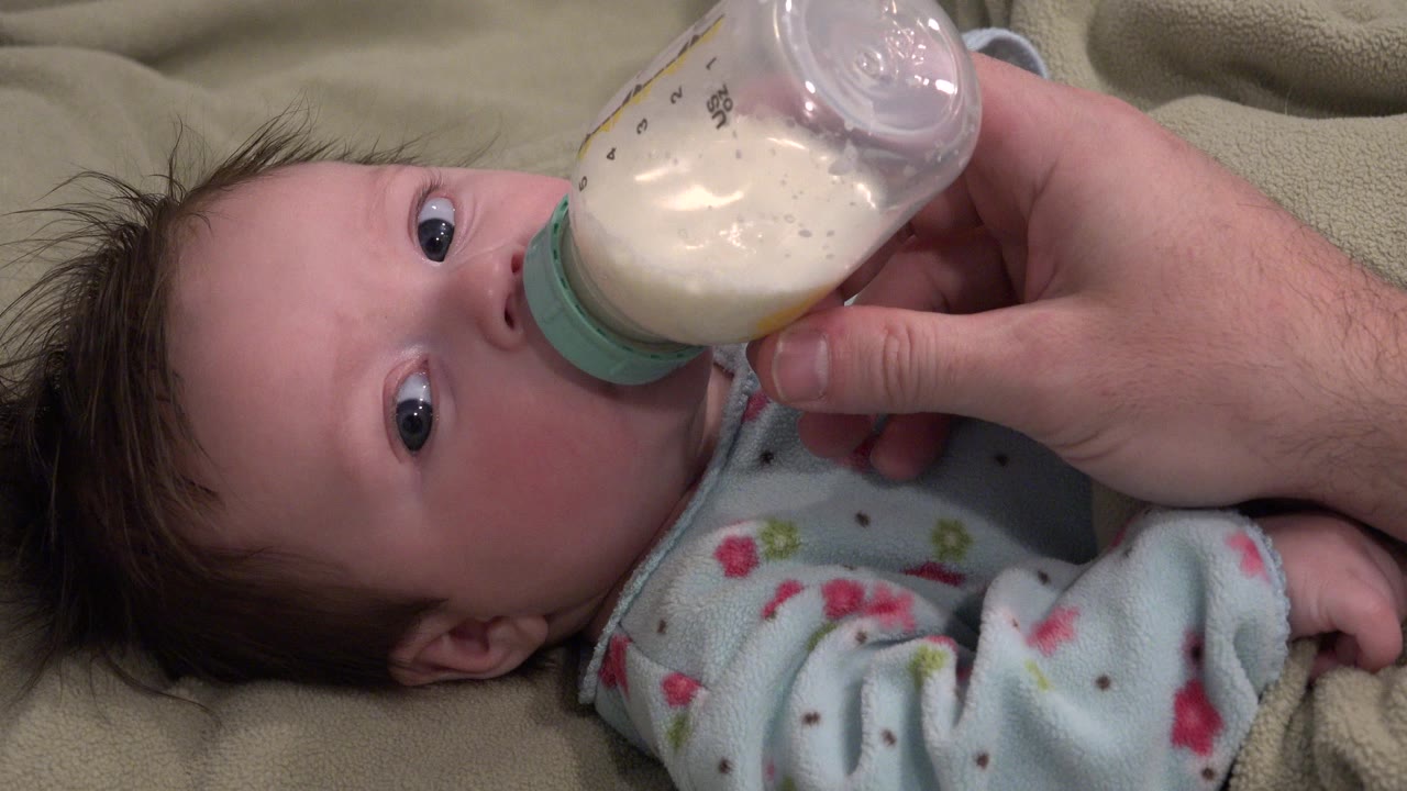 At 0:00, a baby in a pale blue patterned outfit lies on a tan blanket in Westminster, Colorado, while an adult hand holds a milk bottle to the baby's mouth.