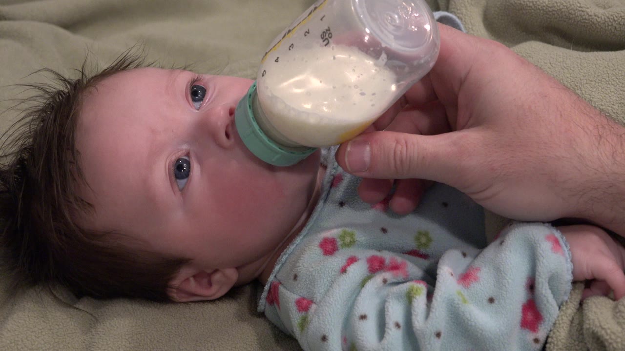 At the start of the Westminster, Colorado clip, a baby in a patterned sleeper lies on a green blanket while an adult hand holds a bottle to the baby's mouth.