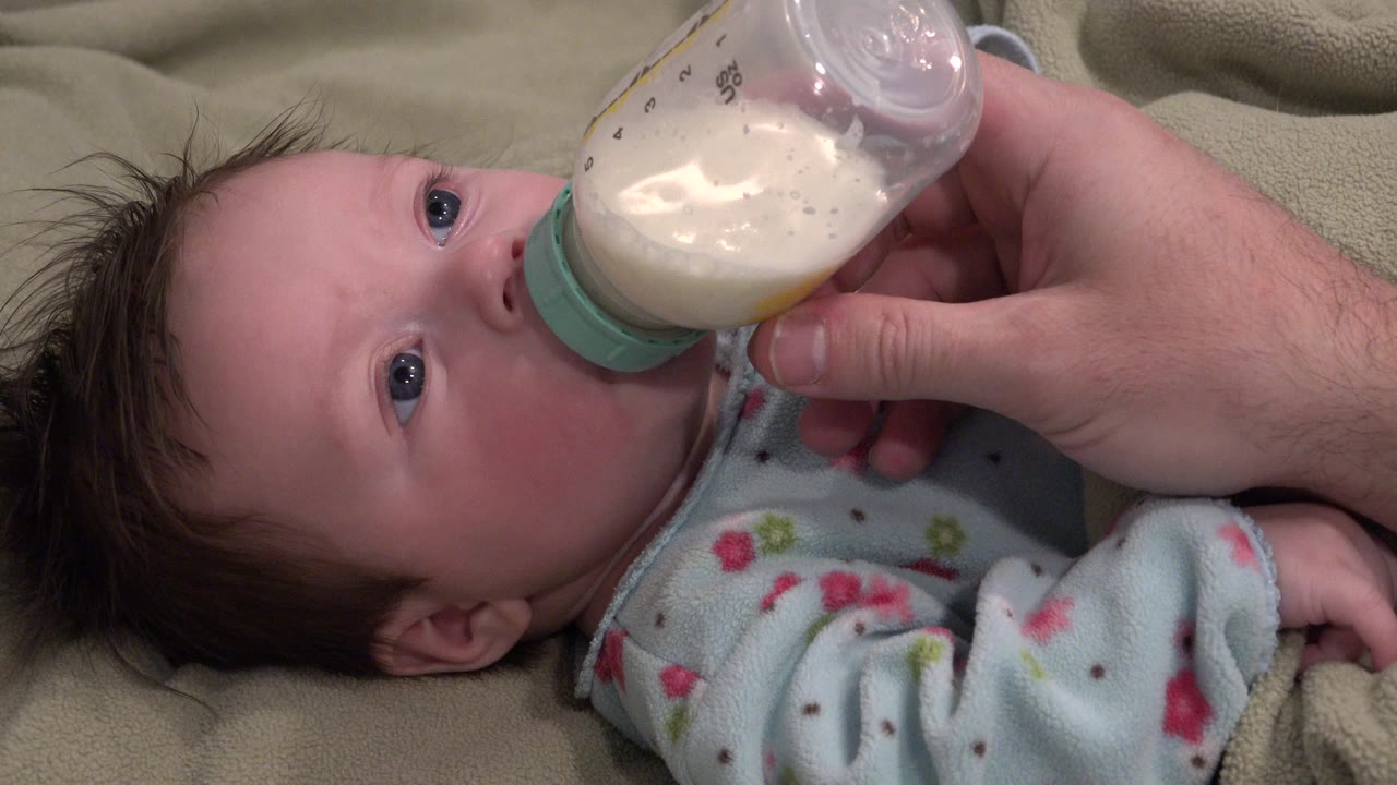 One second in, the baby keeps eyes open toward the bottle as an adult hand steadies the clear bottle against the baby's mouth.
