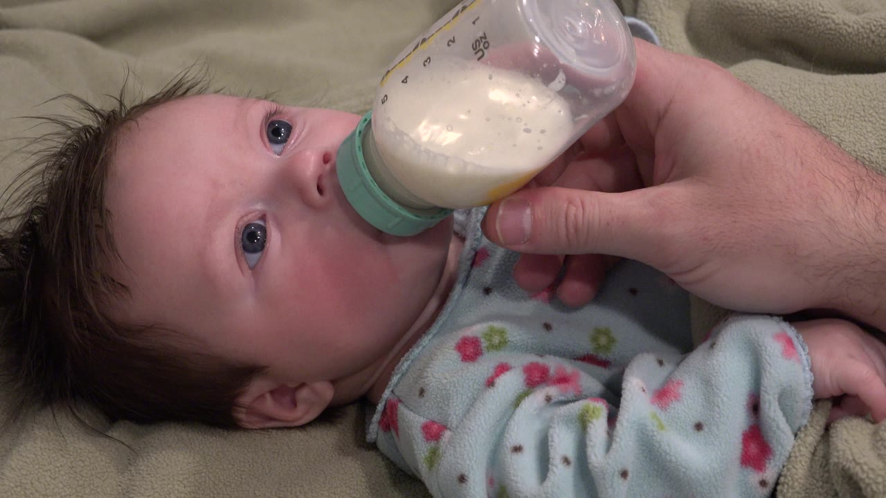 At 0:02, the bottle remains tilted across the baby's face, with milk visible inside and the adult hand holding it from the right side of the frame.