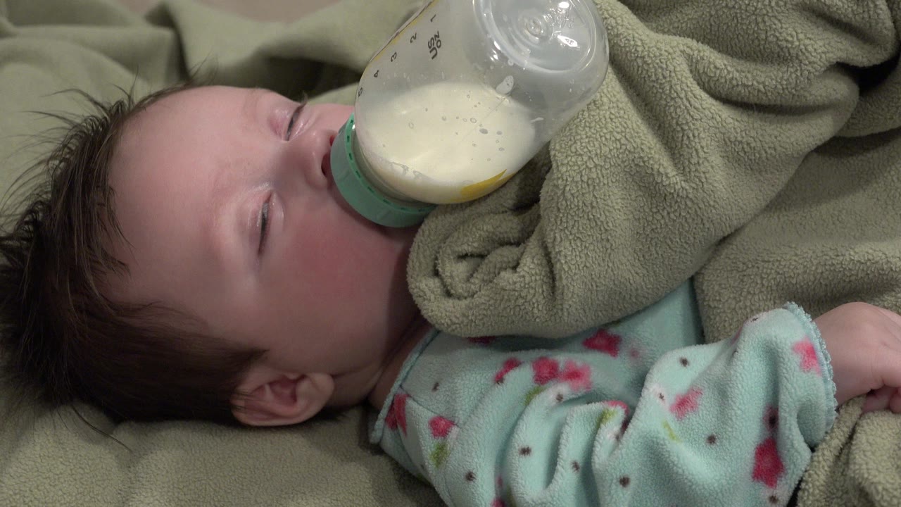 At 0:00 in Westminster, Colorado, a sleepy baby lies on soft blankets with eyes nearly closed while drinking milk from a clear bottle with a green collar.
