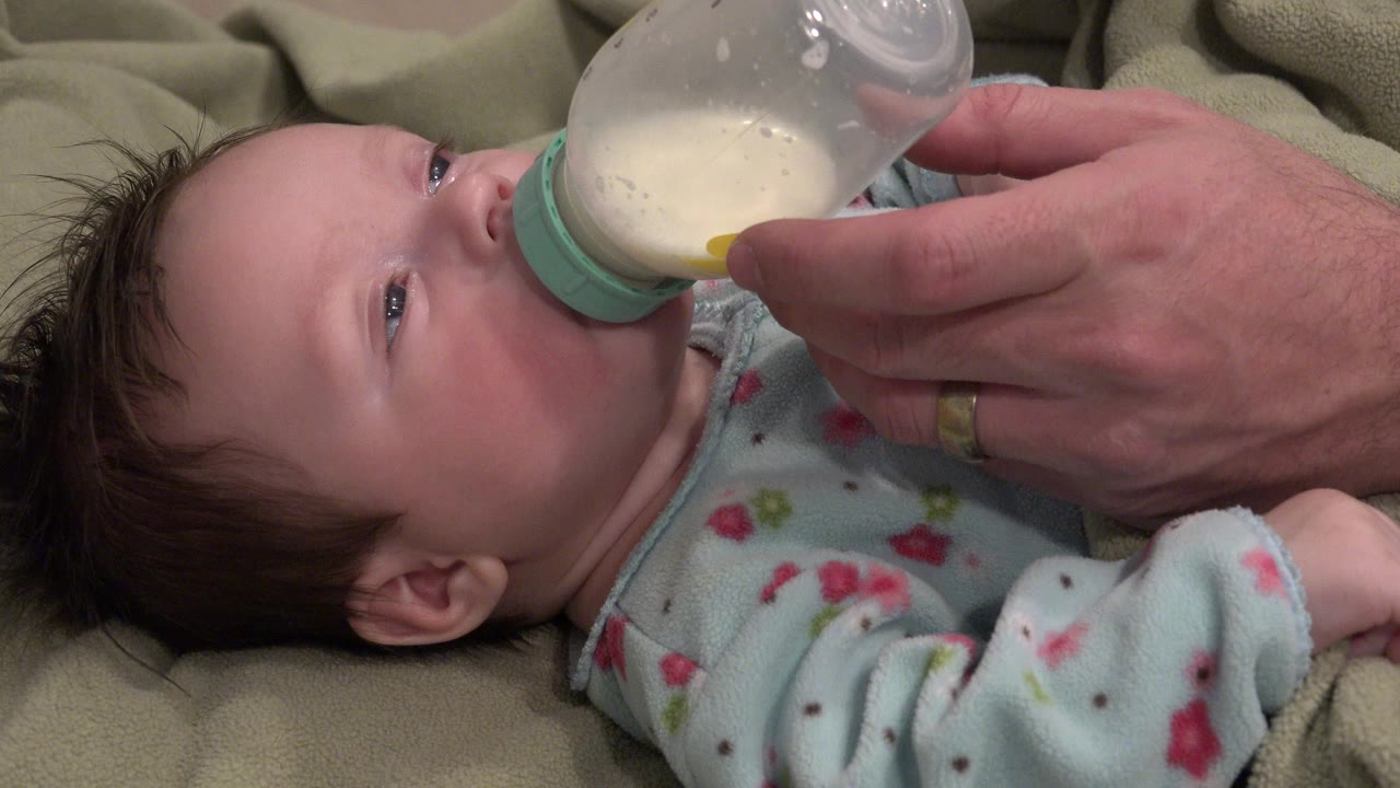 In Westminster, Colorado, a baby lies on beige bedding with eyes open while an adult hand holds a clear bottle to the baby's mouth.