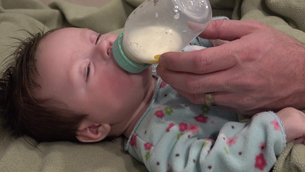 At 0:00 in Westminster, Colorado, a baby lies on a soft blanket as an adult hand holds a partially filled milk bottle to the baby's mouth.