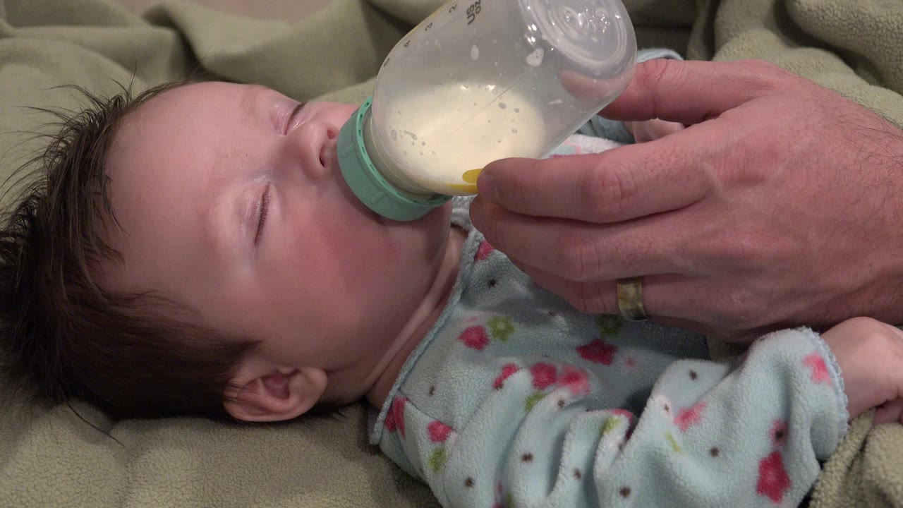 Infant in a blue sleeper drinking from a clear bottle.