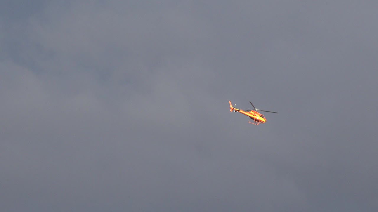 Small yellow-orange helicopter near the upper center of a gray clouded sky in Westminster, Colorado, with no visible people.
