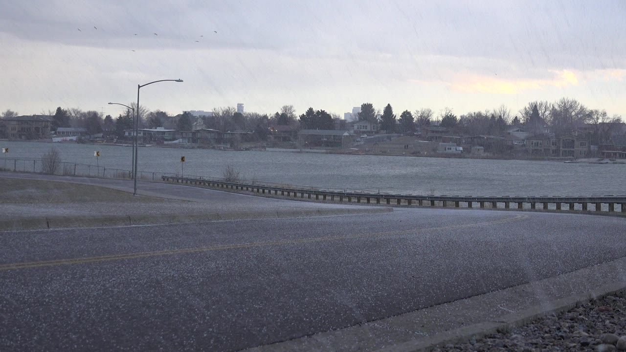 Hail falls over an empty wet street in Westminster, Colorado, with a guardrail, streetlight, fenced water, bare trees, and no visible people.