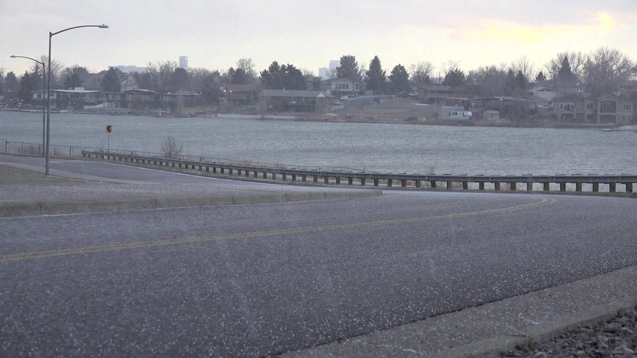 At 0:01 in Westminster, Colorado, the static roadside view holds on the slick pavement, lake, bare trees, and homes across the water, no visible people.
