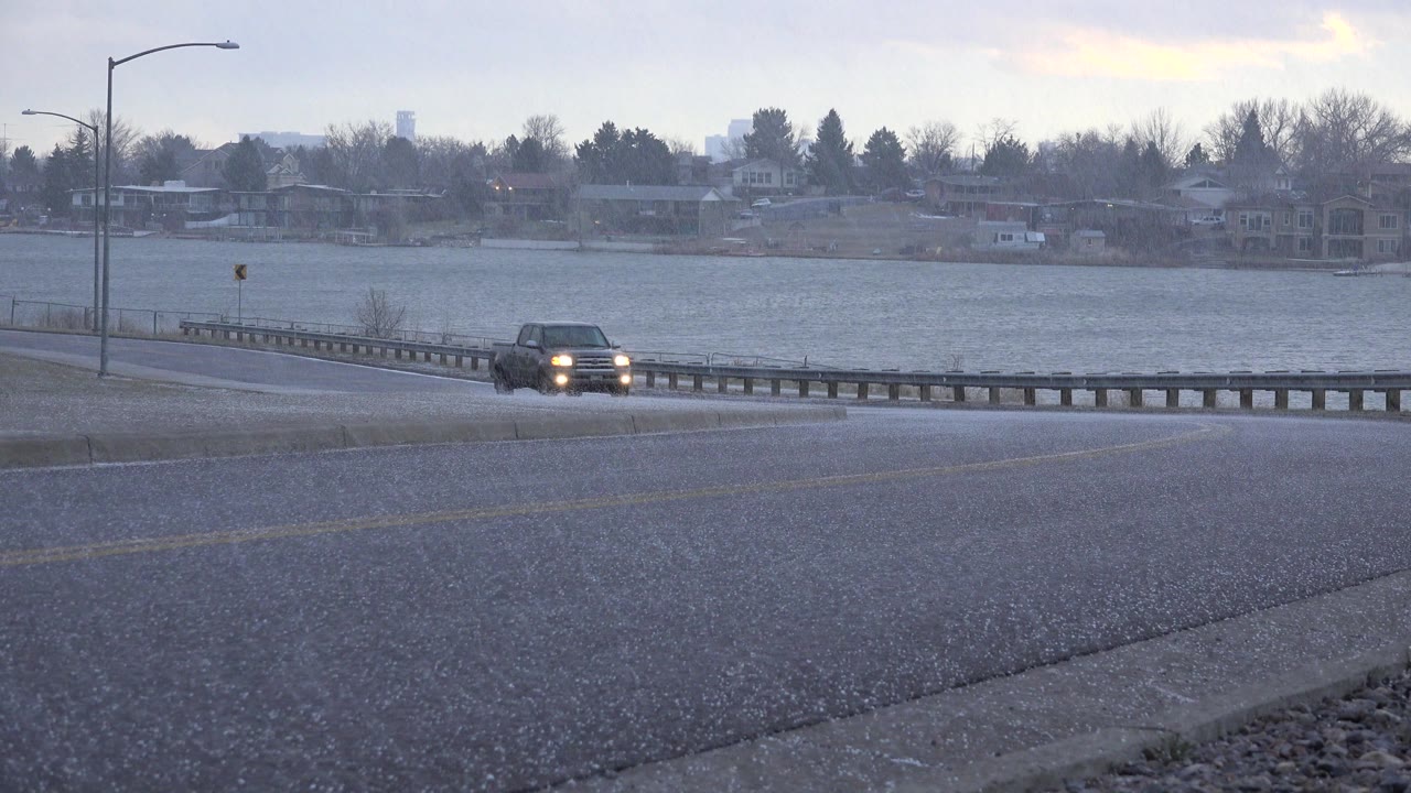 At 0:00 in Westminster, Colorado, a distant dark vehicle with headlights on appears near a wet road, guardrail, lake, streetlight, and background houses, with no visible people.
