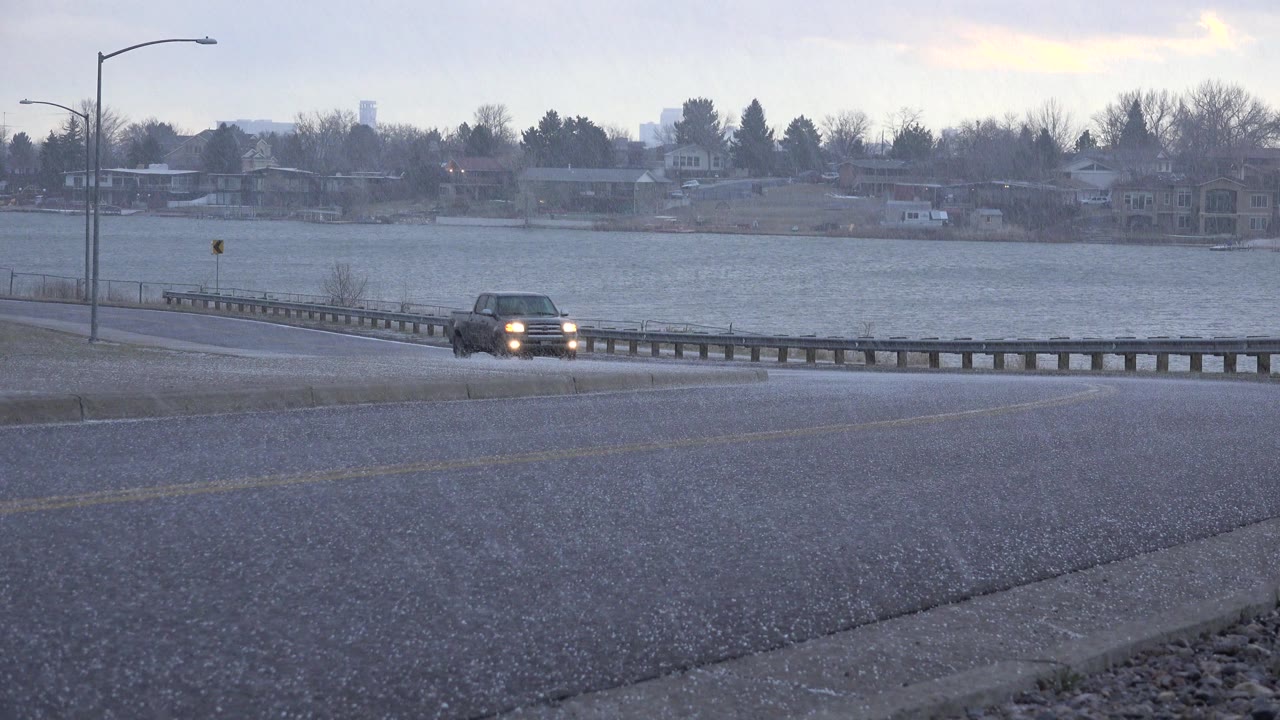 At 0:01 in Westminster, Colorado, the vehicle has moved closer along the wet curve, with its headlights reflecting on the road and the lake and houses behind it, with no visible people.