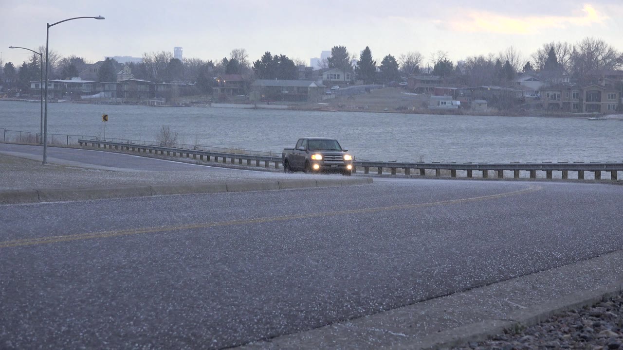 Dark vehicle continues toward camera on a rainy road by a lake.