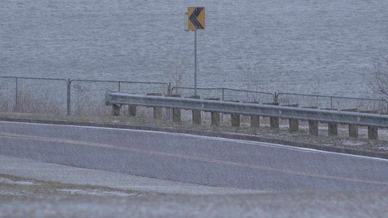 Rainy roadside with guardrail, chevron sign, and water behind it.