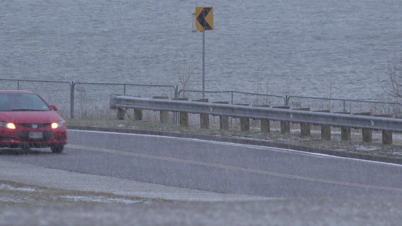 The red car moves farther into the Westminster, Colorado frame on a roadside lane, with headlights lit, a metal guardrail, chain-link fence, muted water, and no visible people.