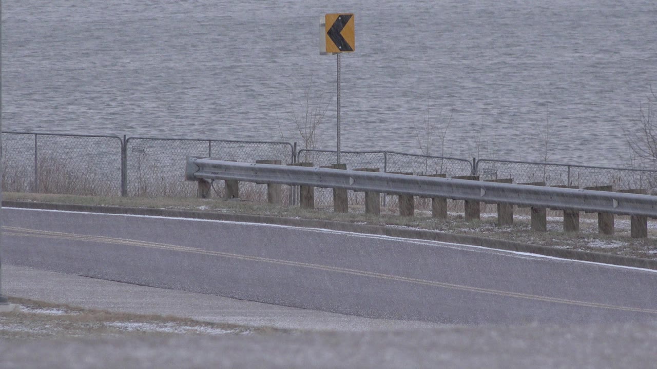 The road remains clear in Westminster, Colorado, with the yellow left-turn chevron sign above the guardrail and rippled gray water filling the background, with no visible people.