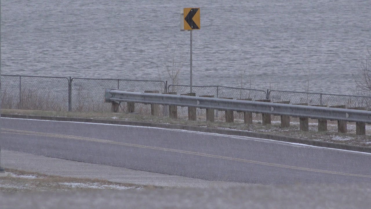 Opening view in Westminster, Colorado shows gray water beyond a chain-link fence, a metal guardrail, a wet roadside edge, and a yellow curve sign, with no visible people.