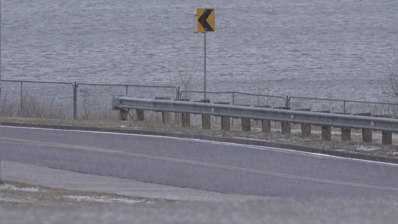 At 0:01, the Westminster, Colorado frame holds on the guardrail and fence line with gray water filling the background and the yellow curve sign near the upper center, with no visible people.