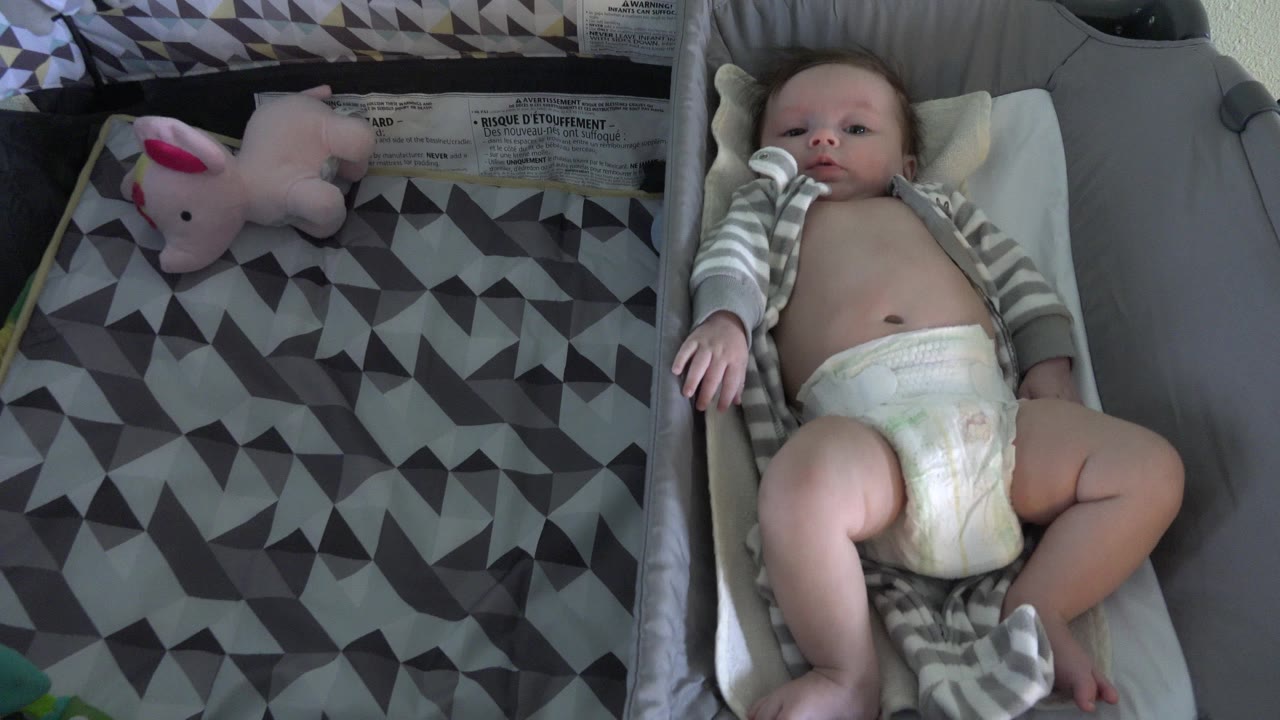 Baby lying in a white bassinet beside a patterned blanket and plush toy.
