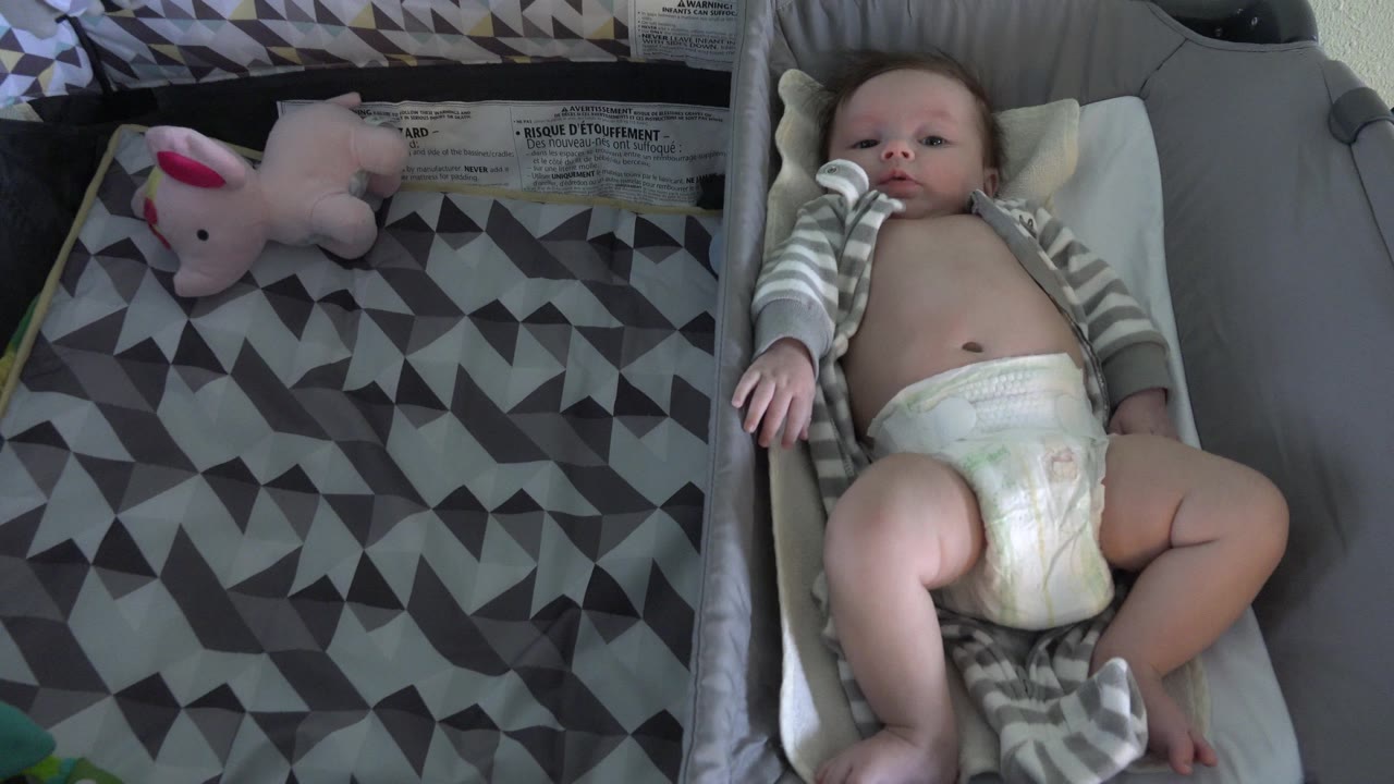 At 0:01, the infant's face is clear as the baby rests in the bassinet, with the striped sleeper open and the patterned blanket still filling the left side.