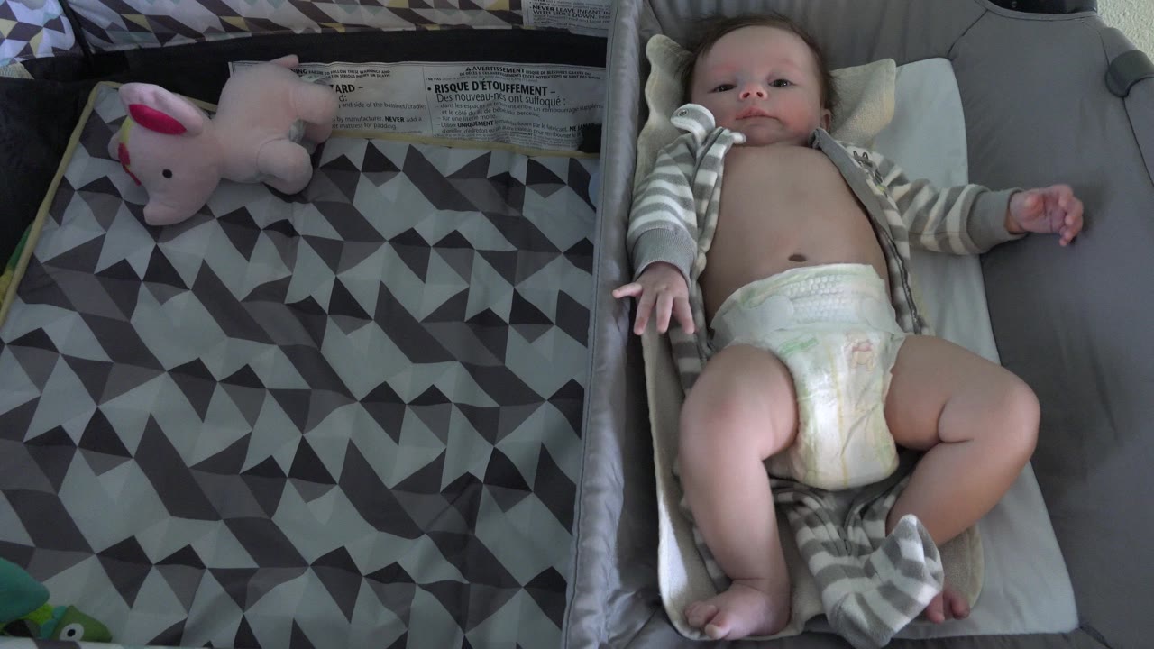 Baby lying on a changing pad beside a gray patterned blanket and pink plush toy.
