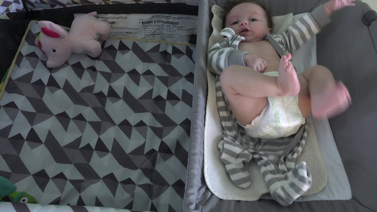 At 0:01 in Westminster, Colorado, a baby lies on a light bassinet pad with legs raised, wearing a diaper and open gray-and-white striped sleeper beside the patterned blanket.