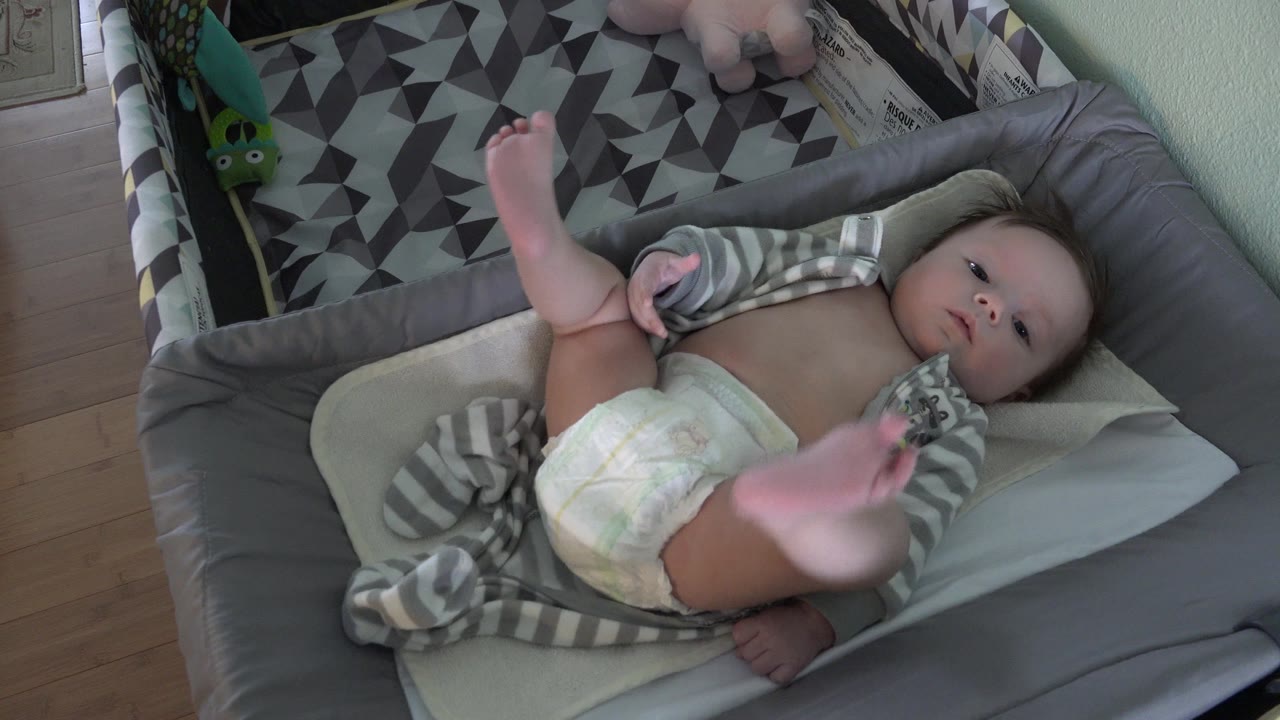 At 0:01, the baby remains on the pale cushion in the gray bassinet, with a blurred foot close to the camera and the patterned bassinet fabric behind the child.