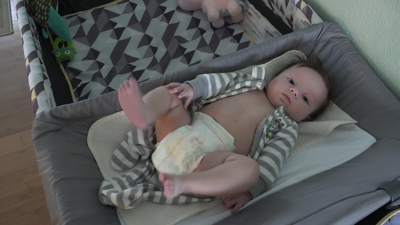 The baby remains on the tan bassinet pad in Westminster, Colorado, with one foot blurred upward from a small kick and the patterned blanket visible behind.