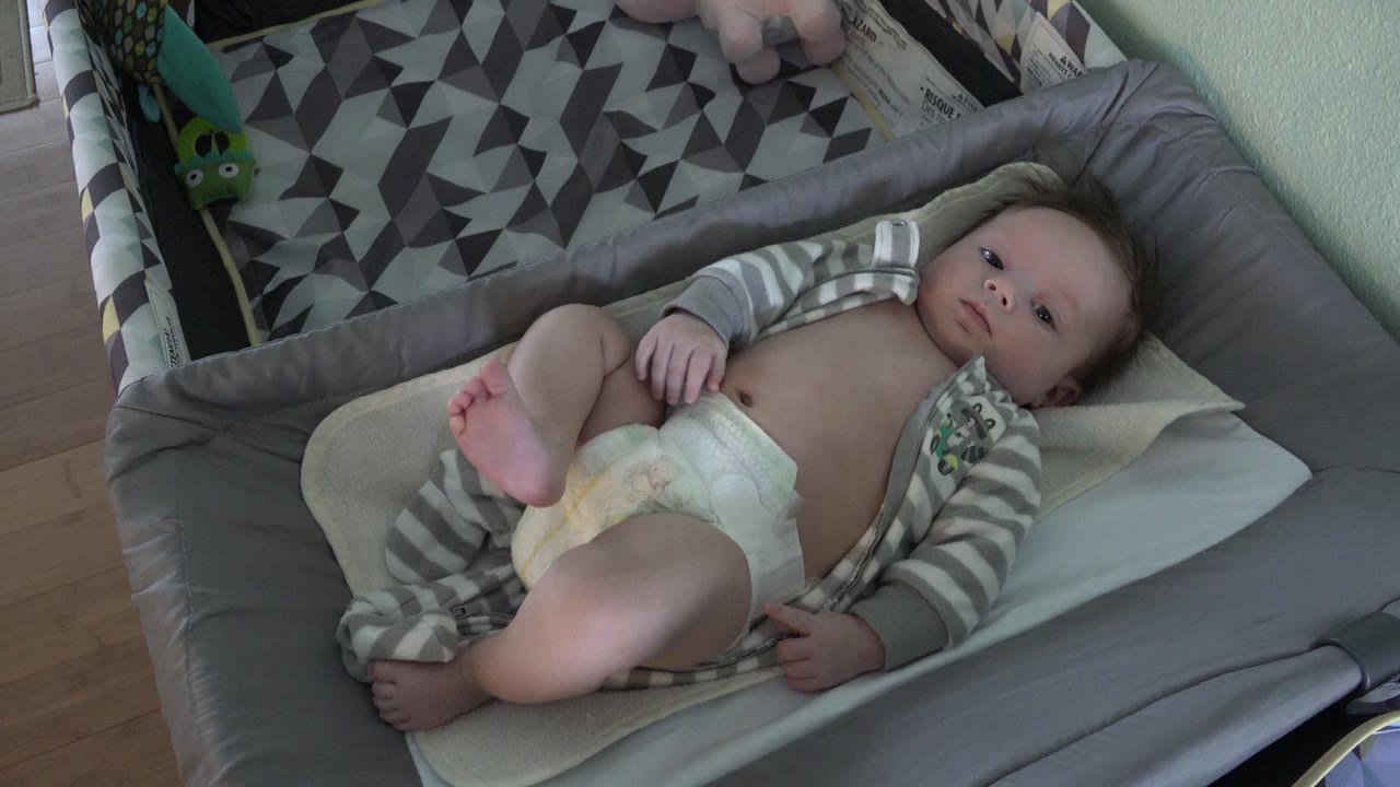 Baby lying in a gray cot with one leg raised.