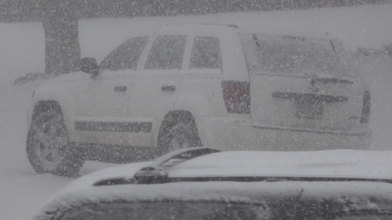 At 0:01, the parked white SUV remains centered in dense snowfall in Westminster, Colorado, with the foreground car roof cutting across the bottom and no visible people.