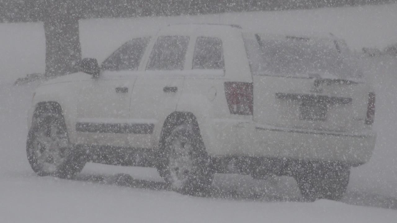 The white SUV remains partly obscured by thick snowfall as another vehicle is visible behind it in Westminster, Colorado, with no visible people.