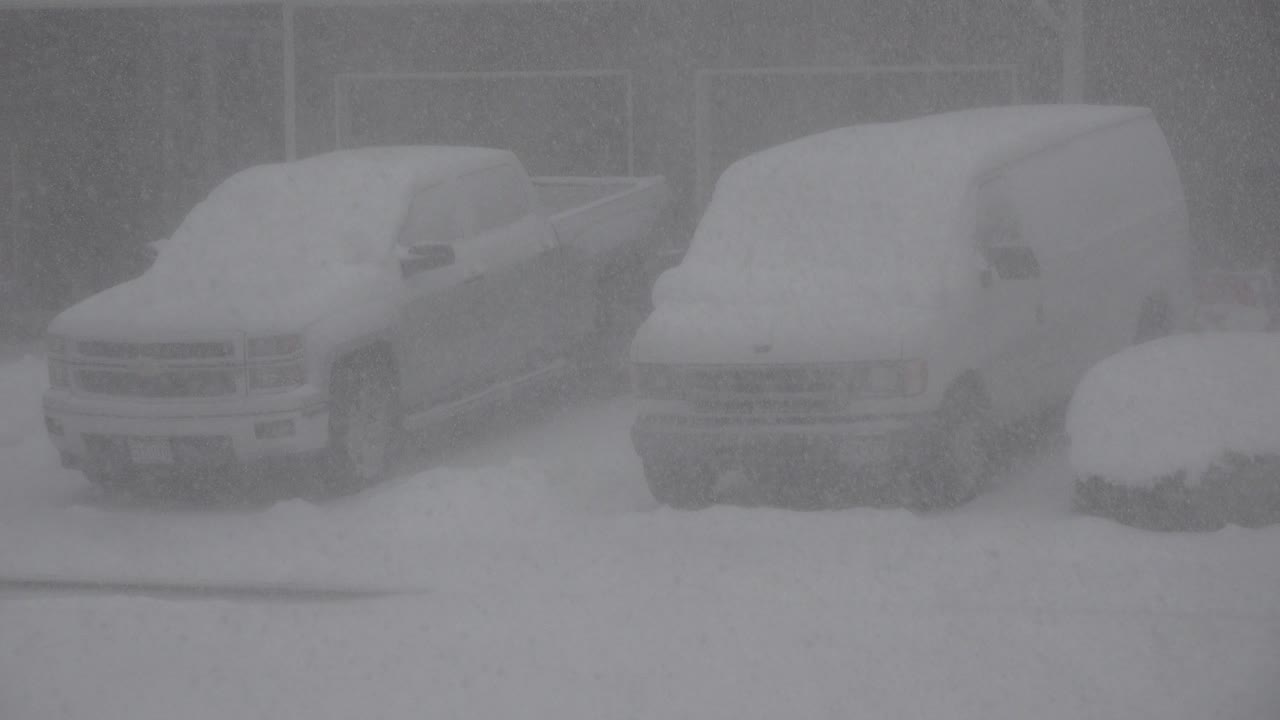 At 0:01 in Westminster, Colorado, heavy snow softens the view of the parked pickup and van, with a rounded snow-covered foreground shape at right and no visible people.