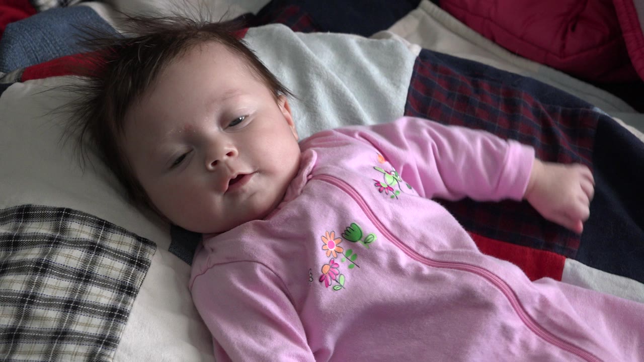 At 0:01, the infant remains on the patchwork quilt in Westminster, Colorado, with one eye partly closed and a small arm extended across dark fabric.