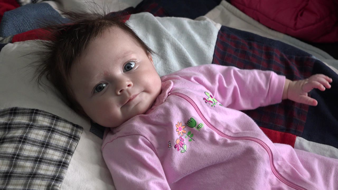 Baby in a pink sleeper lying on a bed and looking at the camera.