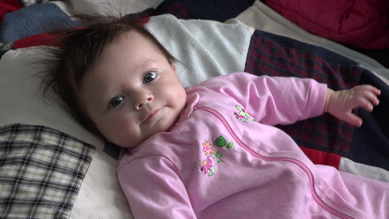 At 0:01, the baby remains on the bed in Westminster, Colorado, looking at the camera while one arm reaches outward over the dark blanket.