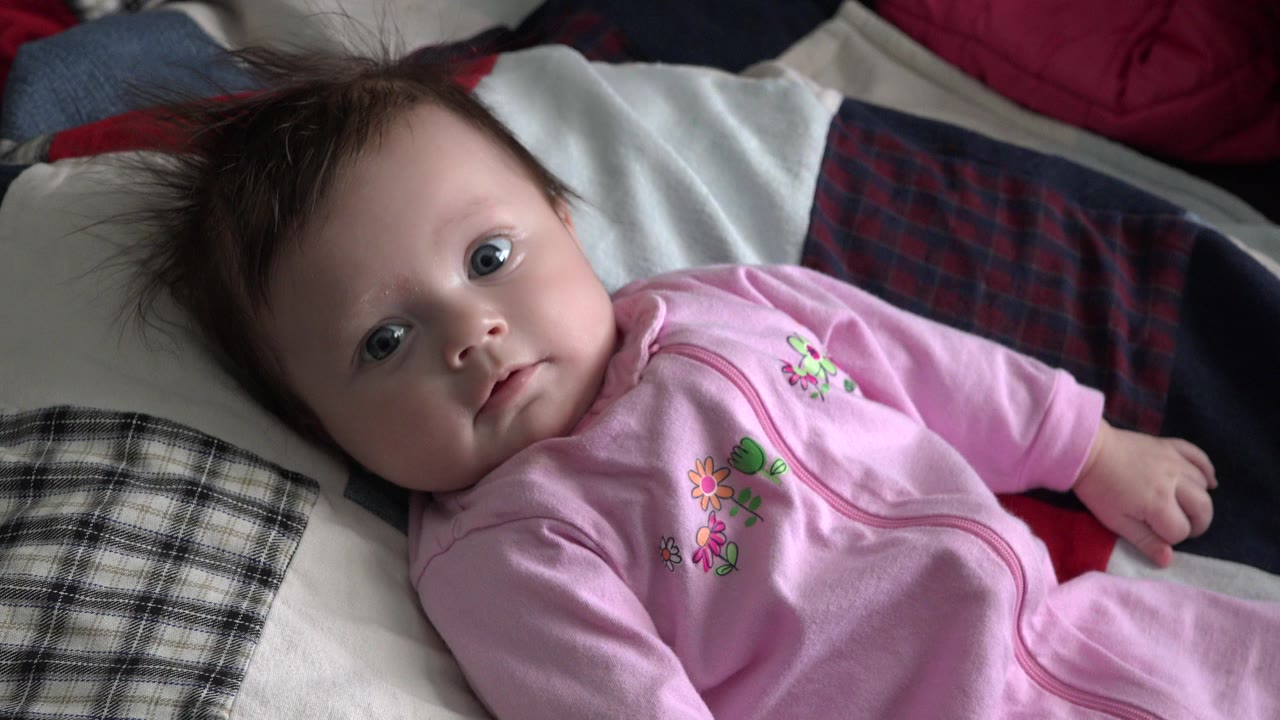The child remains on the patterned bed in Westminster, Colorado, with eyes open toward the camera and a hand resting near the right edge of the frame.