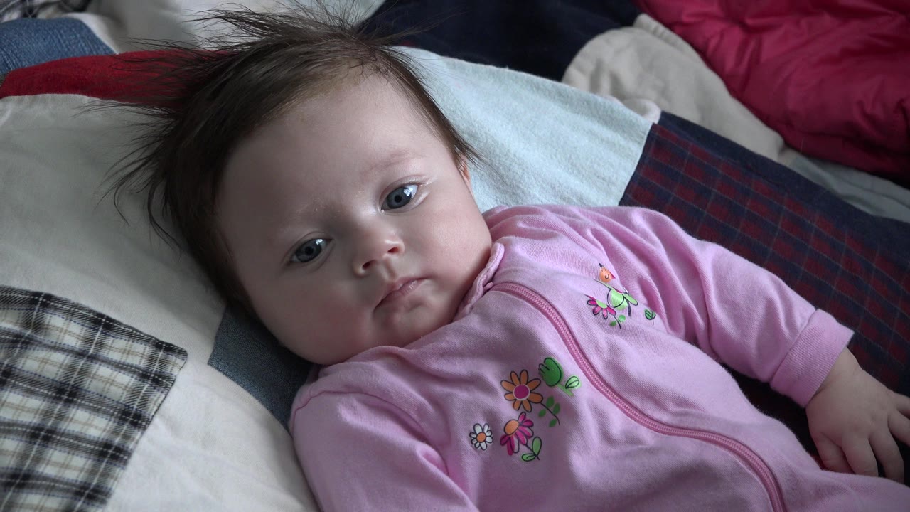 A baby girl in a pink floral sleeper lies on a patterned quilt in a Westminster, Colorado home setting, with pillows and blankets around her.