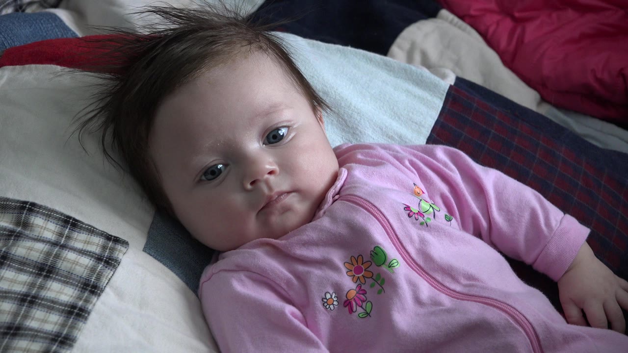 The baby girl remains close to camera on the quilt, her face turned upward with plaid and light-colored pillows behind her in Westminster, Colorado.
