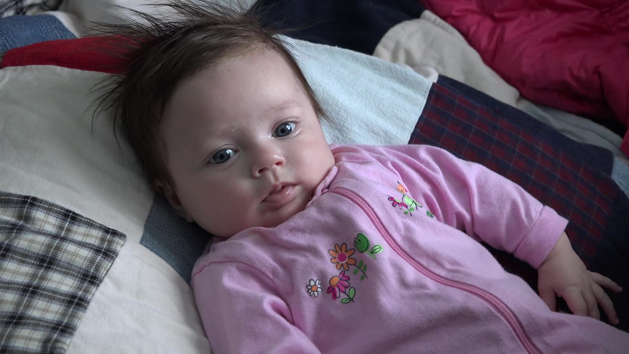 Baby girl lying on bedding with one hand extended.