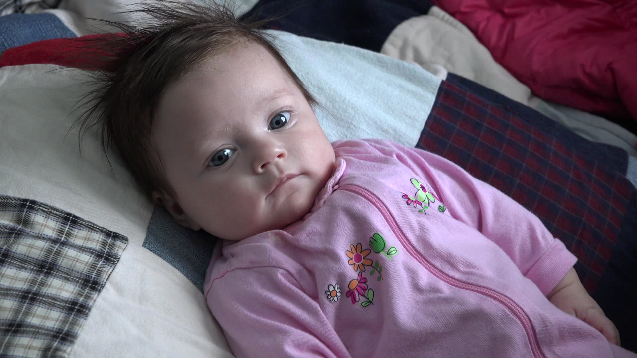 At 0:01, the baby looks toward the camera with the face centered, pink clothing visible, and plaid bedding along the right side of the frame.