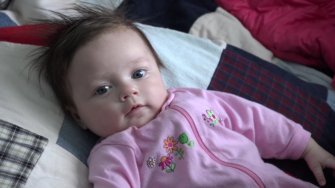 At 0:00, a light-haired baby in a pink flowered romper looks toward the camera while lying on patterned bedding in Westminster, Colorado.