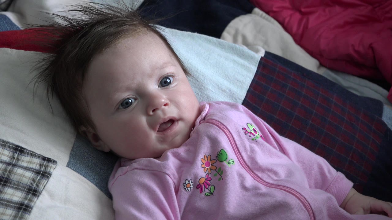 Baby in a pink sleeper lying on a bed and looking toward the camera.