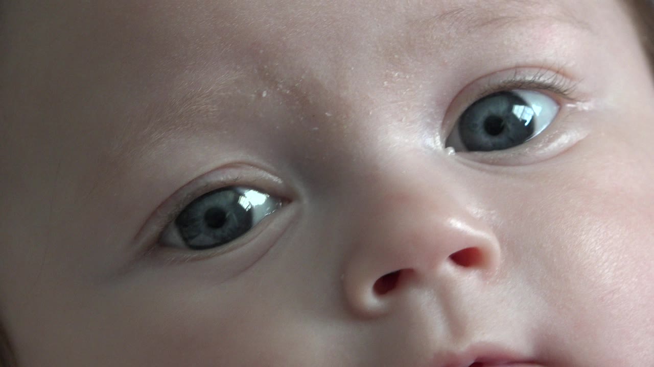 At 0:01, the baby's face remains very close to camera in Westminster, Colorado, with both eyes open, the nose and upper lip partly visible, and faint red hair at the edge.