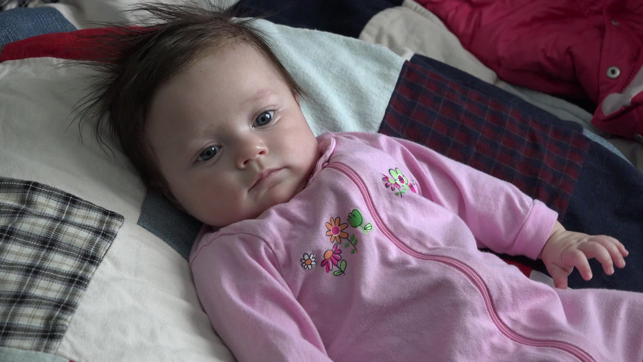 At 0:01, the baby remains on the patchwork quilt in Westminster, Colorado, looking toward the camera with one hand visible near the sleeper zipper.