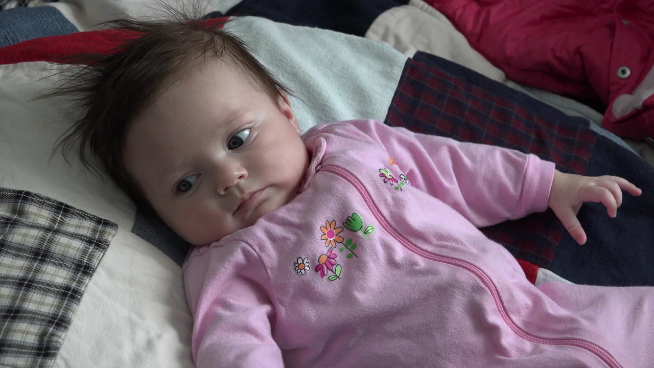 At 0:01, the baby remains reclined on the quilt in Westminster, Colorado, with one arm reaching outward and eyes turned toward the camera area.