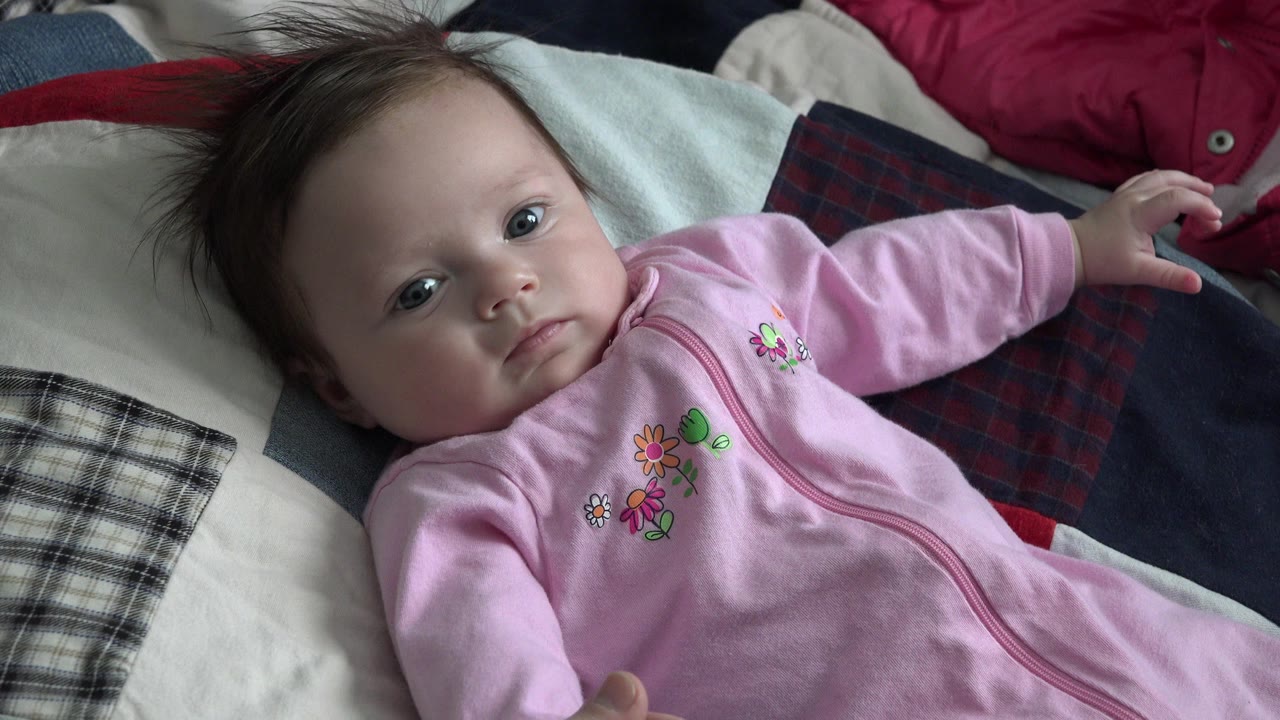 At 0:00, a baby in a pink sleeper lies on a patchwork quilt in Westminster, Colorado, looking up toward the camera with one arm extended.