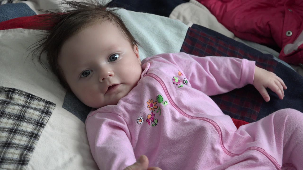 At 0:00, a baby in a pink floral sleeper lies on patterned bedding in Westminster, Colorado, looking toward the camera with one hand near the lower edge.