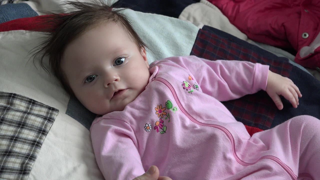 At 0:01, the baby remains centered on the patterned bed in Westminster, Colorado, with the face turned toward the lens and the right arm extended to the side.