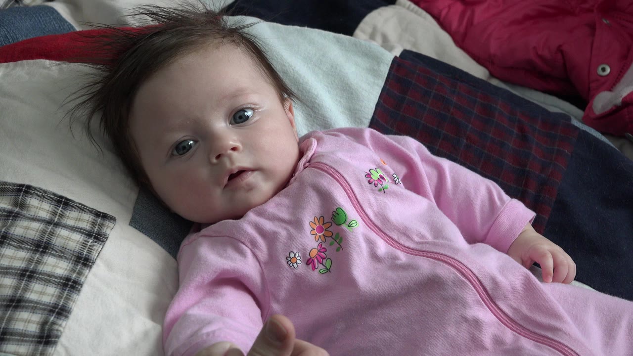 At 0:01, the baby looks toward the camera while lying against plaid pillows and dark bedding in the Westminster, Colorado clip.