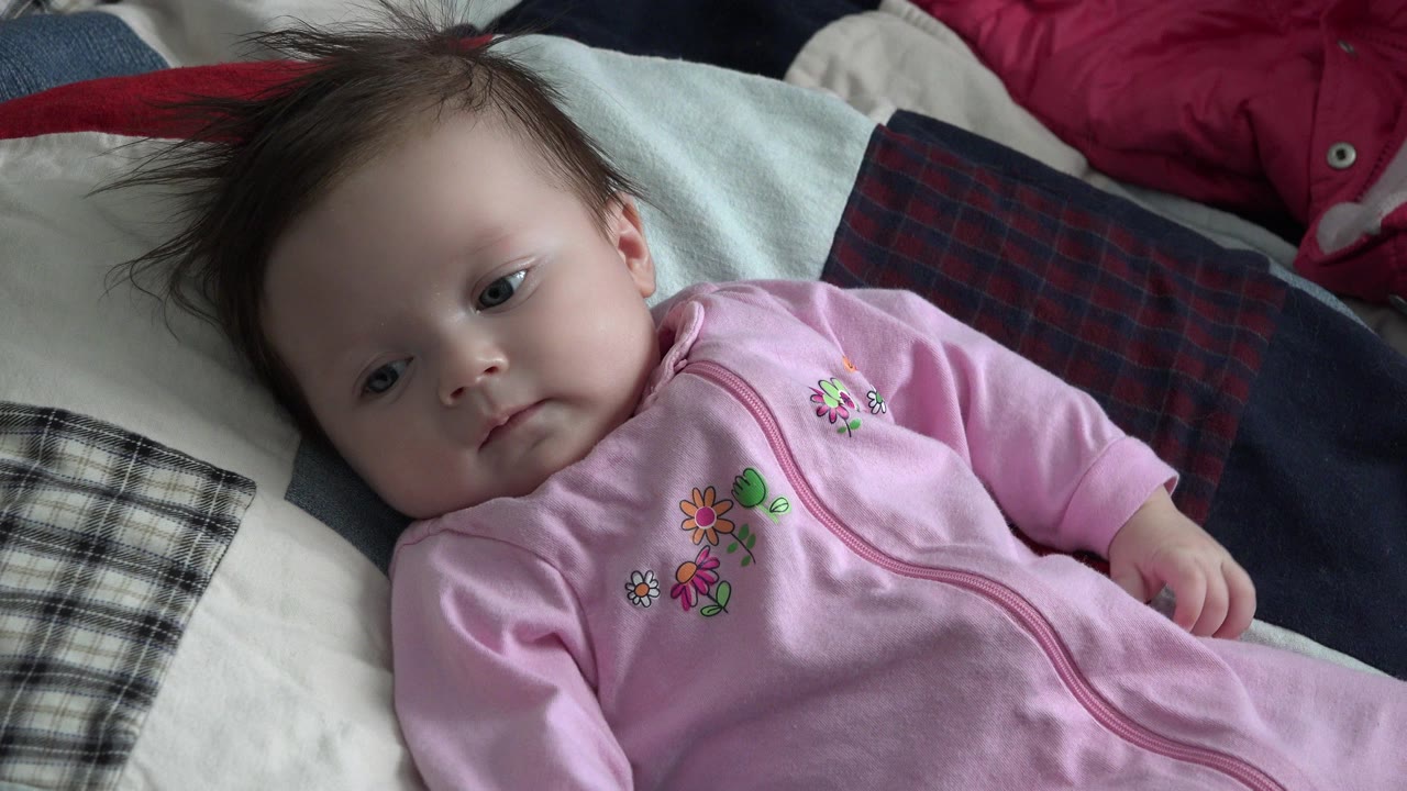 A baby in a pink floral sleeper lies on a patchwork quilt in Westminster, Colorado, looking slightly off camera at the start of the clip.