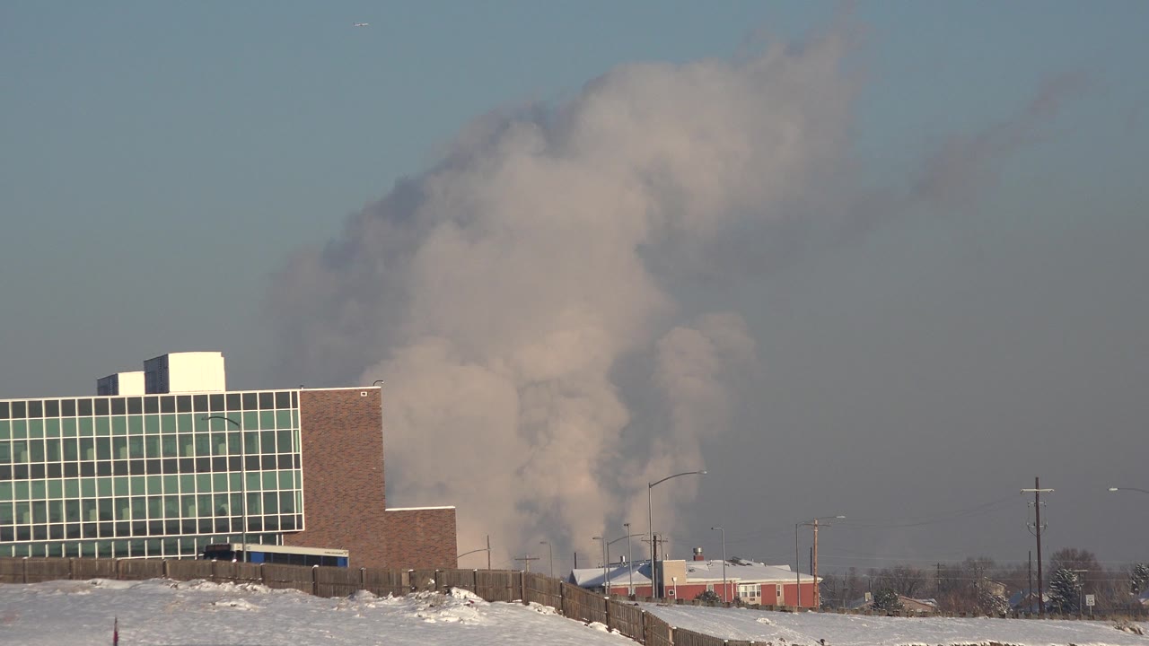 A brick and glass commercial building in Westminster, Colorado stands beside snowy ground as a gray-white plume rises behind it, with no visible people.