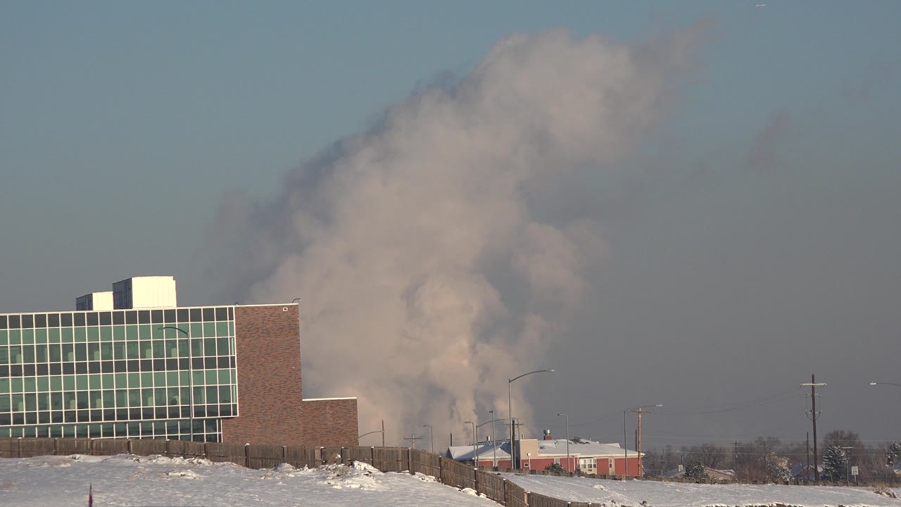 At 0:00 in Westminster, Colorado, a brick and glass building stands beside snow-covered ground as a large white cloud rises behind low urban structures, with no visible people.
