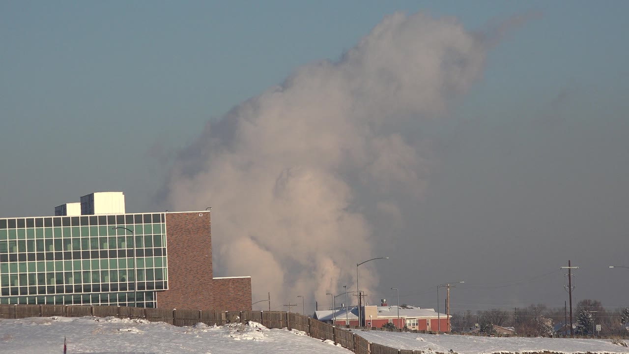 At 0:00 in Westminster, Colorado, a brick and glass industrial building stands beside snow-covered ground as a gray smoke plume rises behind it, with no visible people.